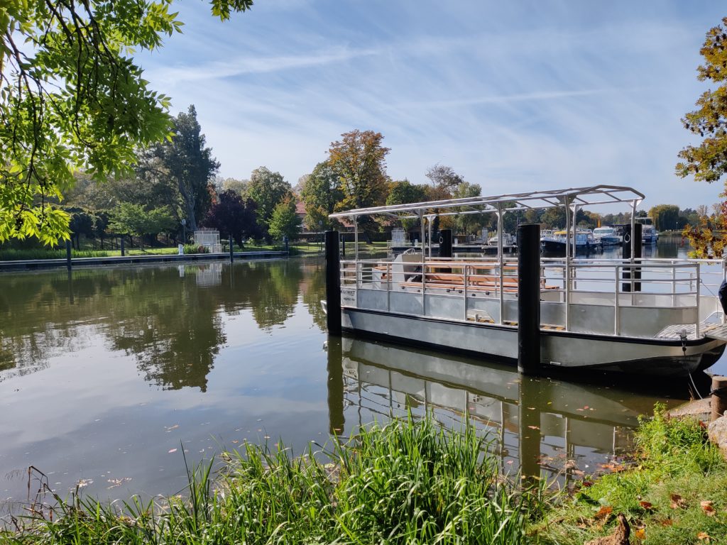 Bateau 100% électrique à quai, dans l'eau en Moselle. La navette est sur l'eau et est entourée de verdure.